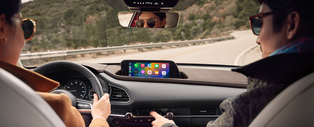 Deux personnes conduisant un Mazda CX-5 sur une route sinueuse, avec l'écran central affichant les applications de connectivité. Un paysage boisé est visible à travers le pare-brise.