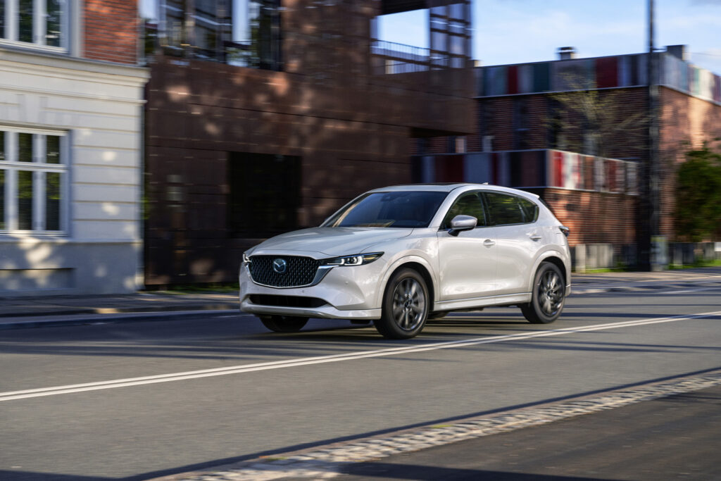 Un Mazda CX-5 blanc conduit dans une rue résidentielle bordée d'immeubles en briques.